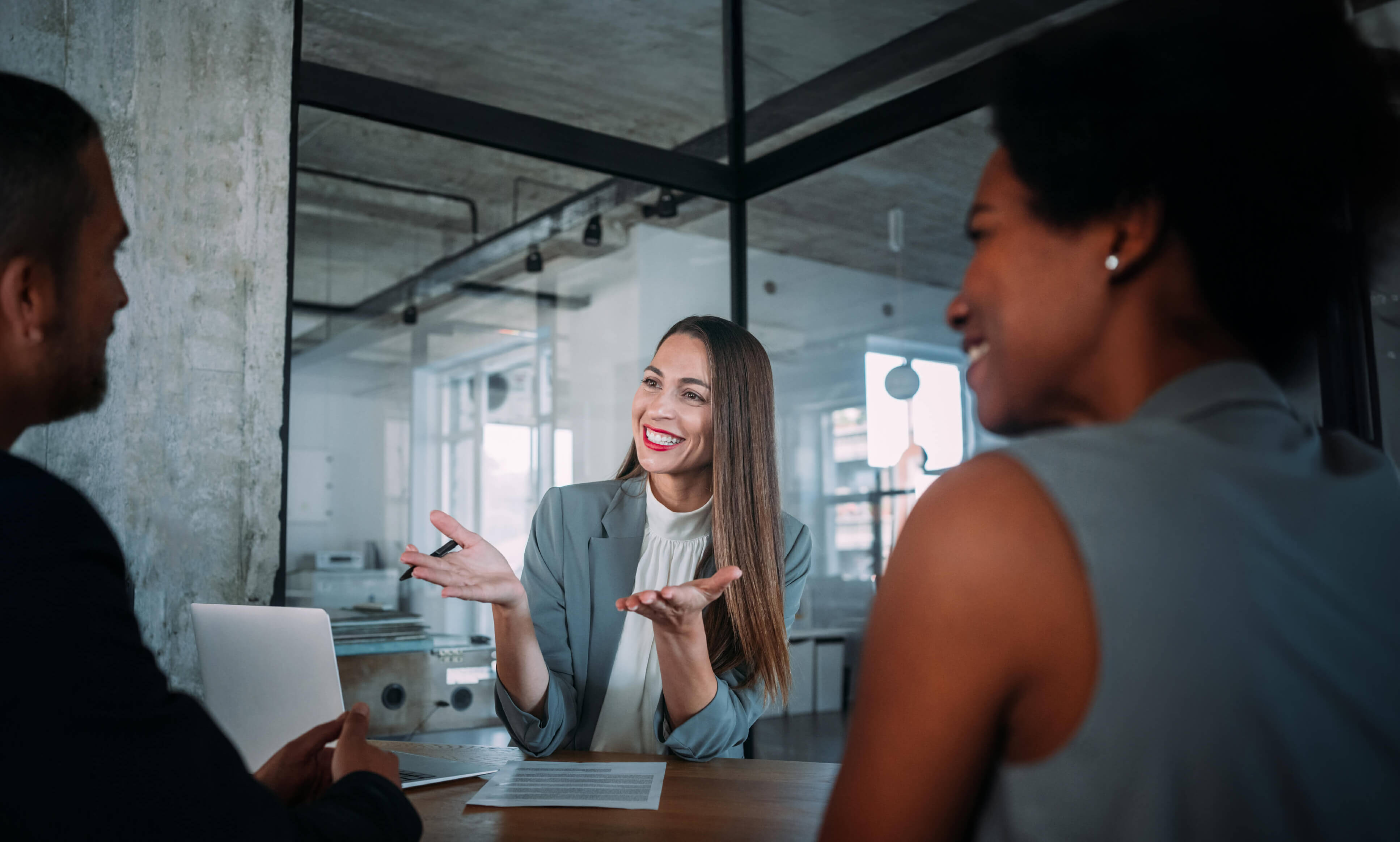 Angeregte Beratungssituation einer jungen, lächelnden Beraterin mit zwei weiteren Personen, die ihr in einem modernen Loft-Büro gegenübersitzen. Marina Adler – Marketing mit Weitblick